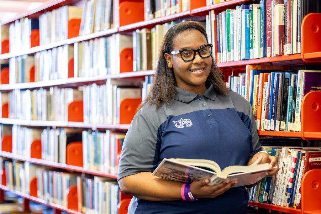 戴着黑框眼镜的女学生拿着一本打开的书, 微笑着站在图书馆的书架前